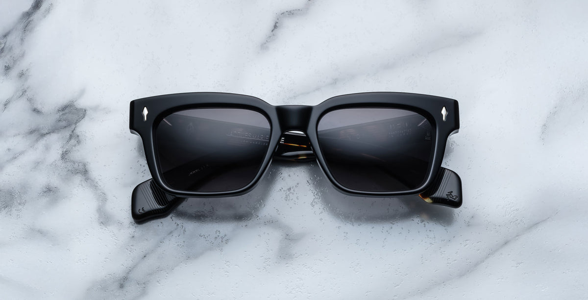Jacques Marie Mage Molino in Noir 11 are black rectangular sunglasses with thick frames and dark lenses, resting on a white marble surface. The curved arms feature a subtle design detail near the hinges.