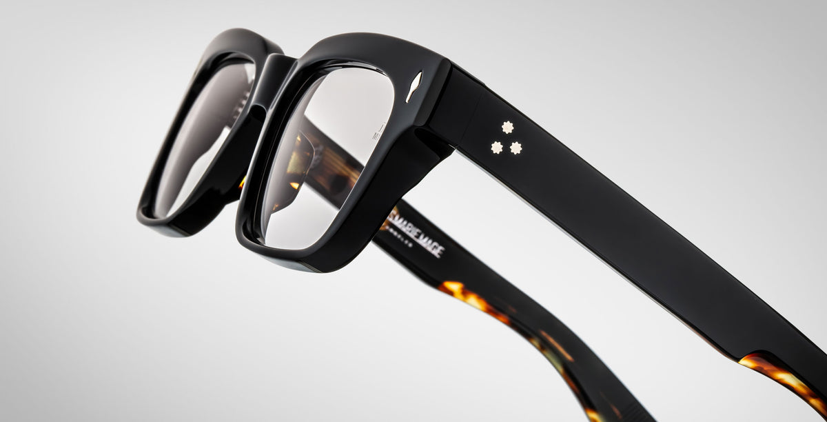Close-up of the Jacques Marie Mage Molino by Jacques Marie Mage, showcasing stylish thick black frames with silver dot accents on the arms and tortoiseshell inner details, set against a light gradient background.