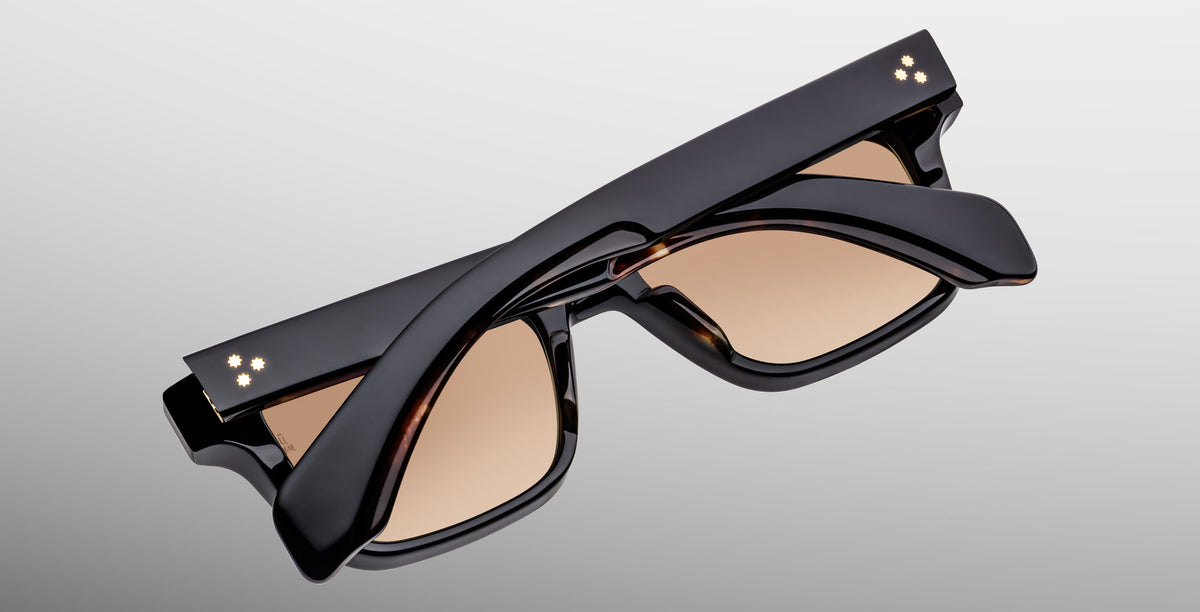 The Jacques Marie Mage Molino sunglasses in Noir 15 feature a stylish black frame with brown-tinted lenses and small decorative studs on the arm hinges, elegantly folded on a light gray background.