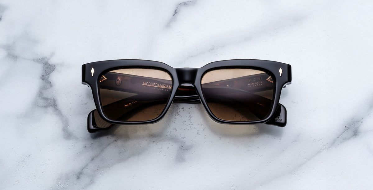 The Jacques Marie Mage Molino in Noir 15 black square-framed sunglasses with brown-tinted lenses and gold-accented arms, rests on a white marble surface; subtle branding appears inside the temples.