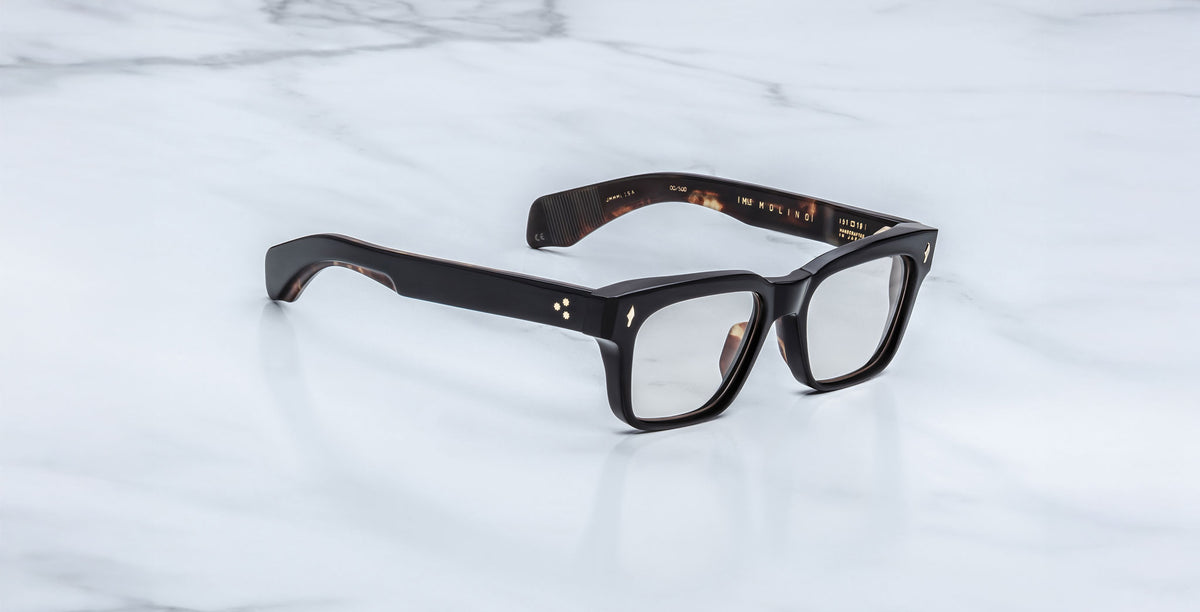 The Jacques Marie Mage Molino eyeglasses in Noir 15 feature bold, black rectangular frames and are displayed on a white marble surface.