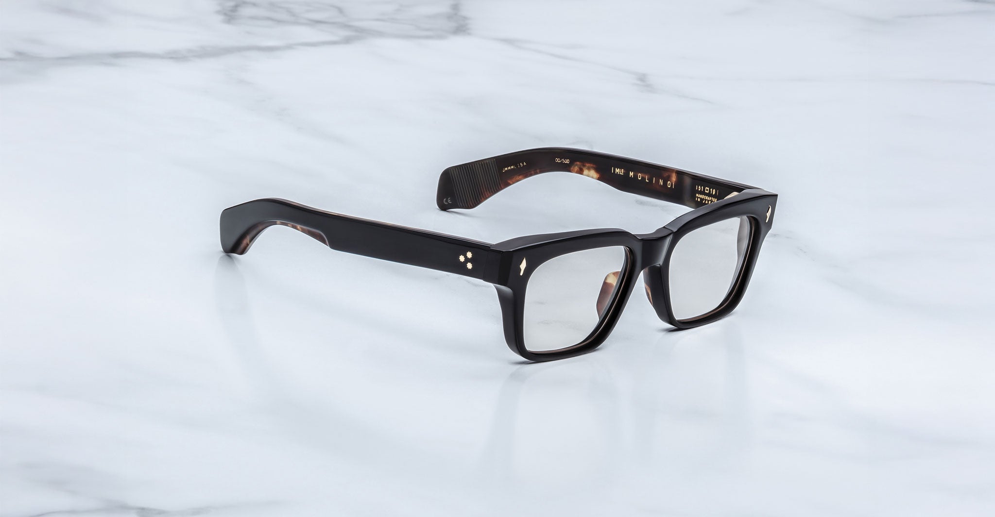 The Jacques Marie Mage Molino eyeglasses by COMING SOON feature bold, black rectangular frames and are displayed on a white marble surface.