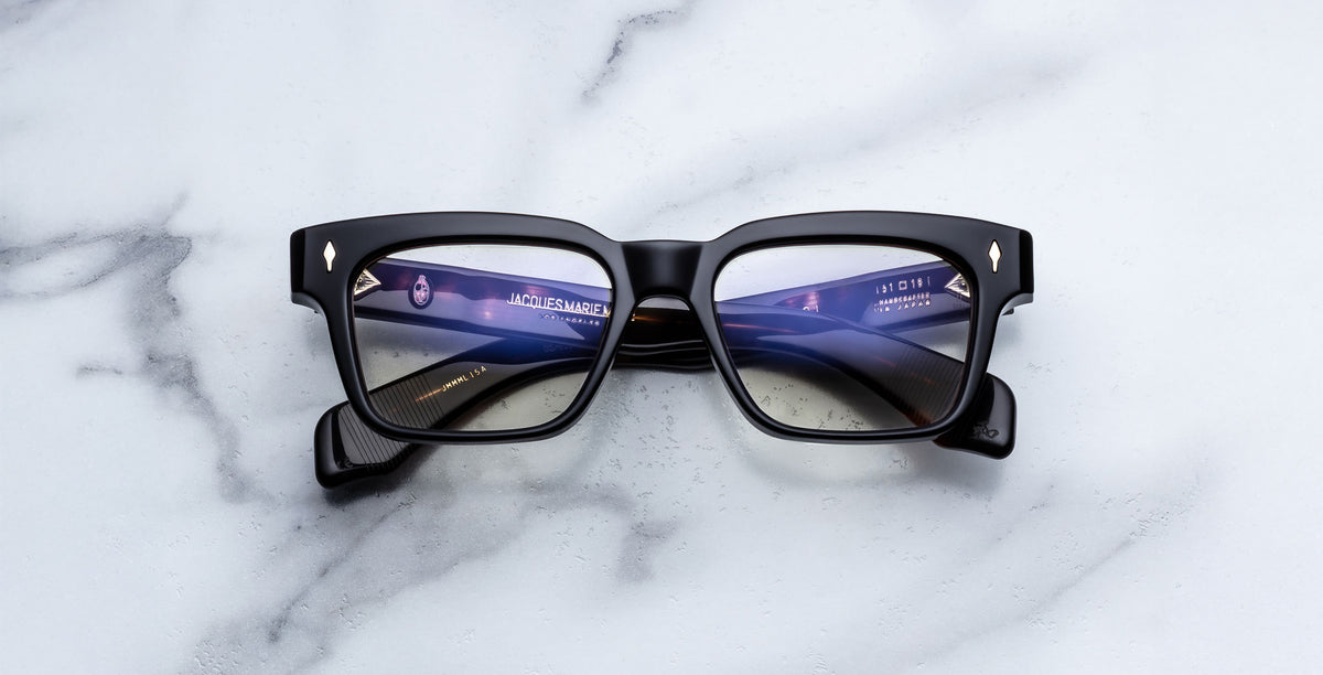 The Jacques Marie Mage Molino in Noir 15 features black square frames and blue-tinted lenses, shown resting on a white marble surface with screen reflections visible on the lenses.