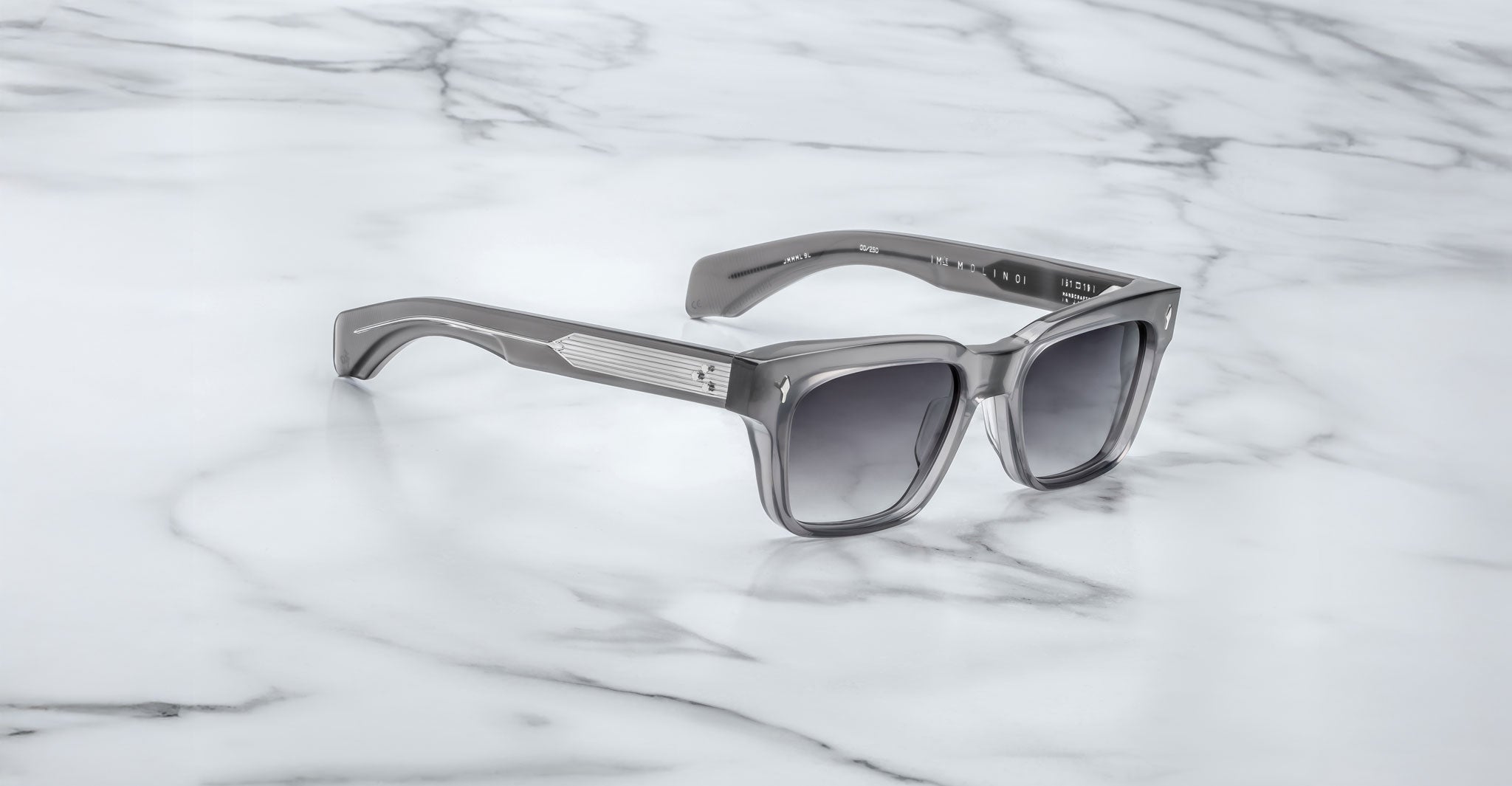The Jacques Marie Mage Molino by COMING SOON, featuring silver frames and dark lenses, rests on a white marble surface with gray veining.