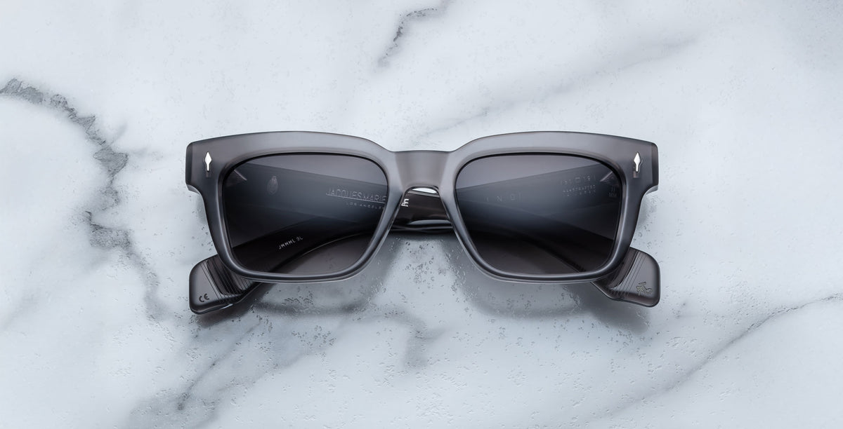 Jacques Marie Mage Molino rectangular gray sunglasses in Prime, featuring dark tinted lenses, rest on a white marble surface.