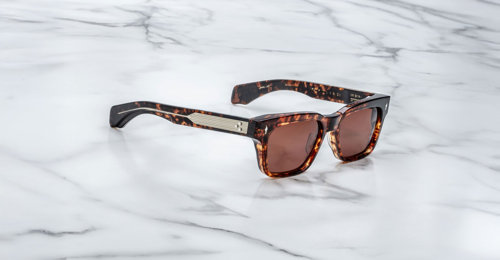 The Jacques Marie Mage Molino are tortoiseshell sunglasses with rectangular brown lenses, wide arms, and a metallic accent by the hinges, displayed on a white marble surface.