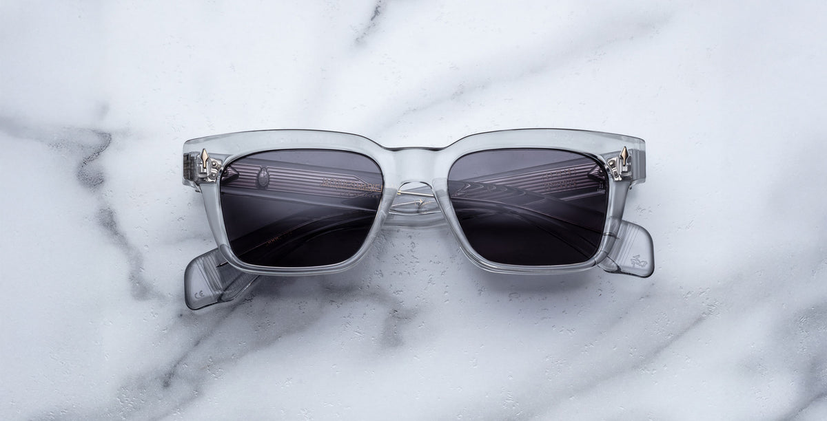 A pair of Jacques Marie Mage Molino square, translucent gray sunglasses with dark lenses rests on white marble, reflecting a ceiling light pattern.