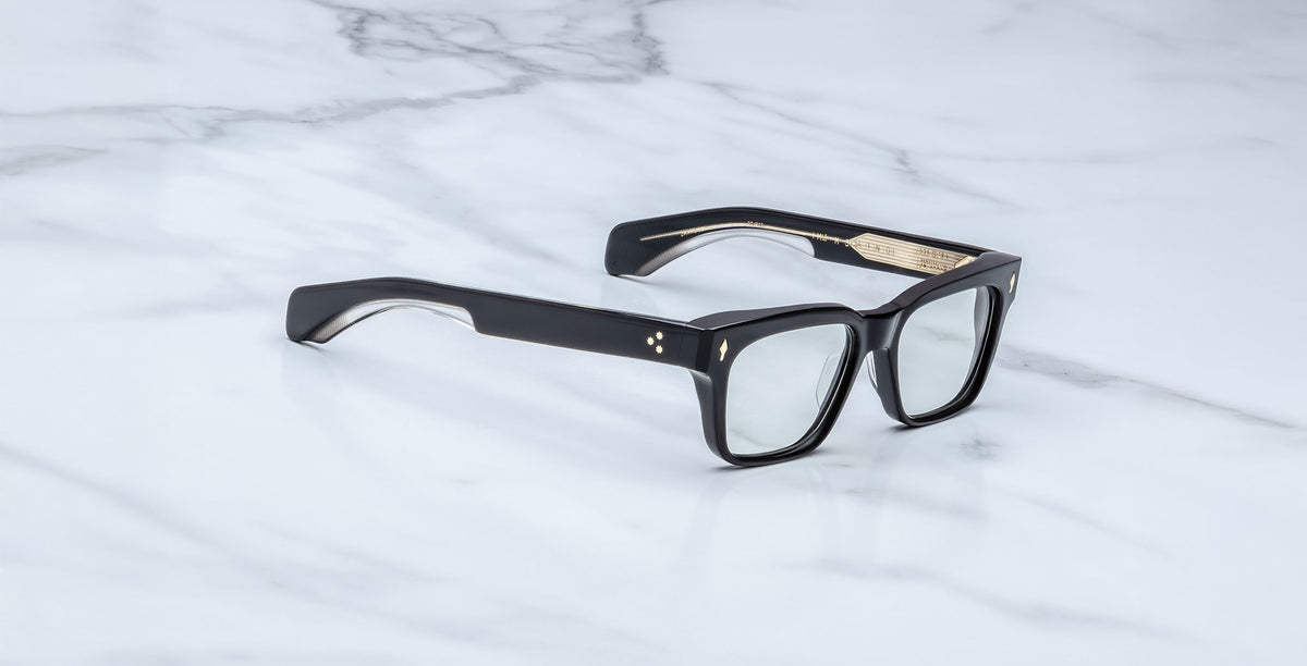 The Jacques Marie Mage Molino featuring black rectangular thick frames and clear lenses, rests on a white marble surface.