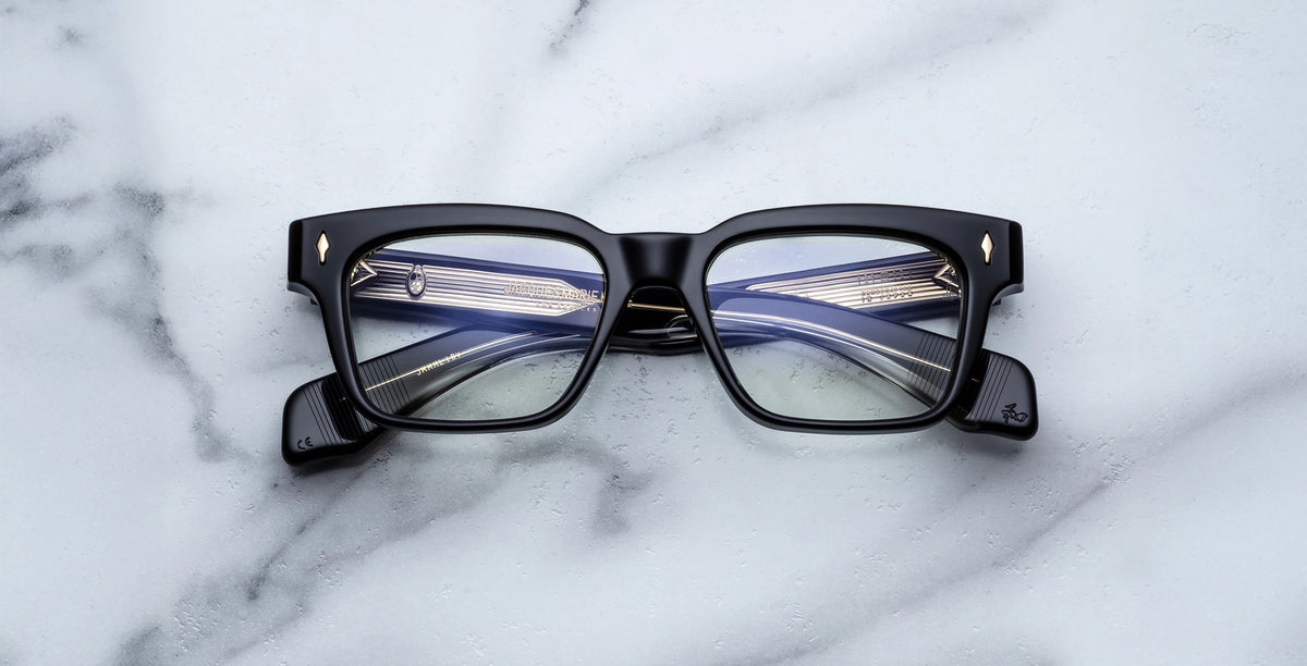 The Jacques Marie Mage Molino Venom, black rectangular eyeglasses with thick frames, rest on a white marble surface, their lenses reflecting the temples and hinges.
