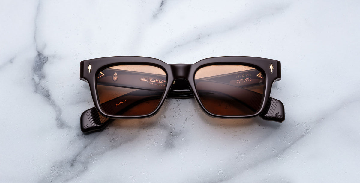 A pair of Jacques Marie Mage Molino square sunglasses with black frames and brown-tinted lenses is displayed face-up on a white marble surface.