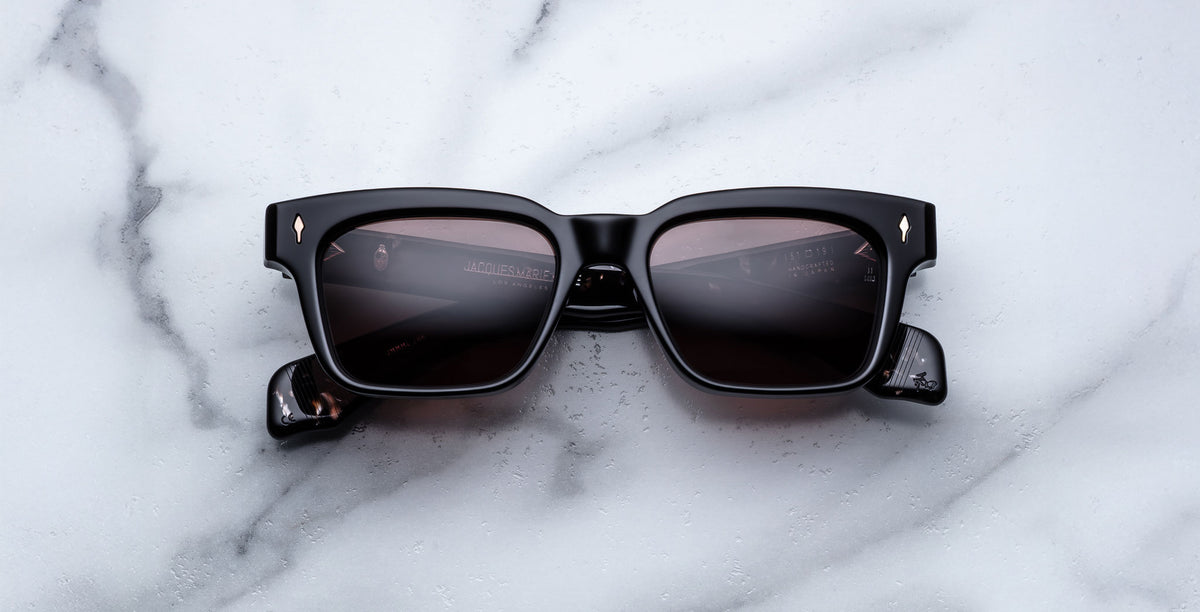 Jacques Marie Mage Molino black rectangular sunglasses with dark tinted lenses and gold diamond-shaped accents on the upper corners, displayed on a white marble surface.