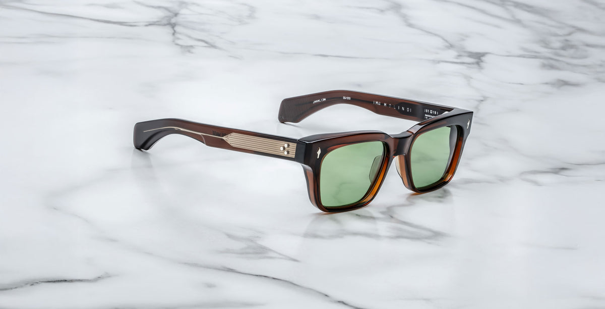 The Jacques Marie Mage Molino sunglasses feature a stylish square brown frame with green lenses and metallic temple accents, displayed on a white marble surface.