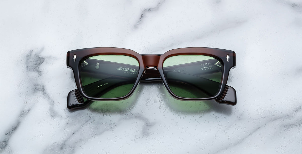 The Jacques Marie Mage Molino sunglasses in Zoltar feature a brown and black rectangular frame with green-tinted lenses, showcased on a white marble surface.