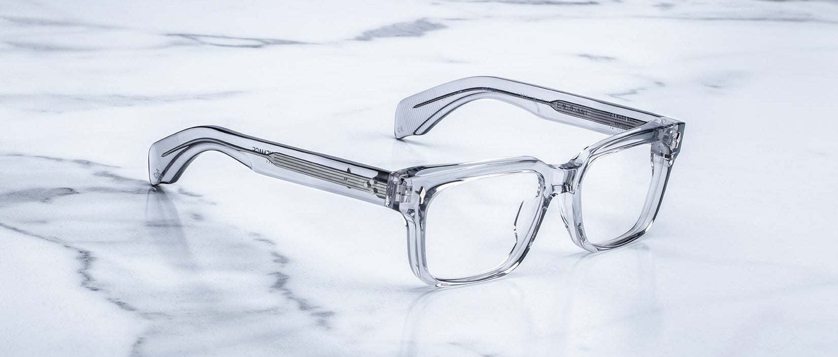 The Jacques Marie Mage Molino 55 features clear, rectangular eyeglasses with thick transparent frames, showcased on a white marble surface with subtle gray veining.