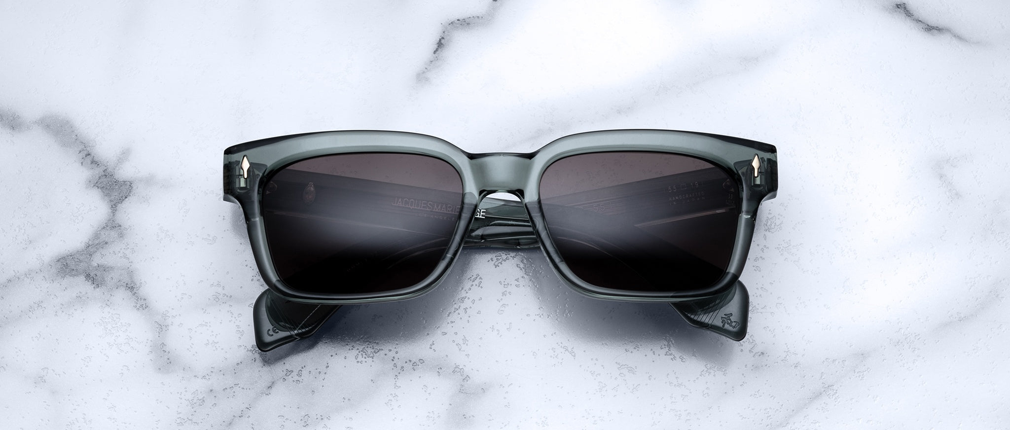 A pair of Jacques Marie Mage Molino 55 square-framed sunglasses with dark lenses rests on a white marble surface.