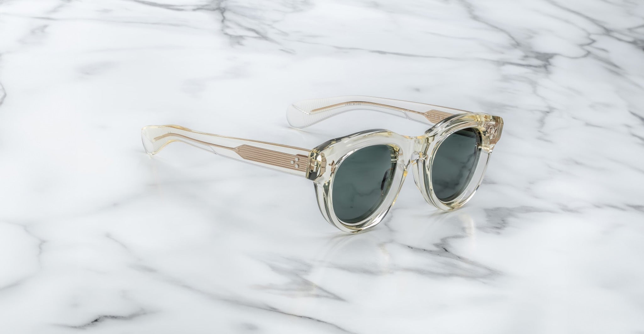 Jacques Marie Mage Molitor sunglasses with clear frames and dark lenses displayed on a white marble surface featuring gray veining.