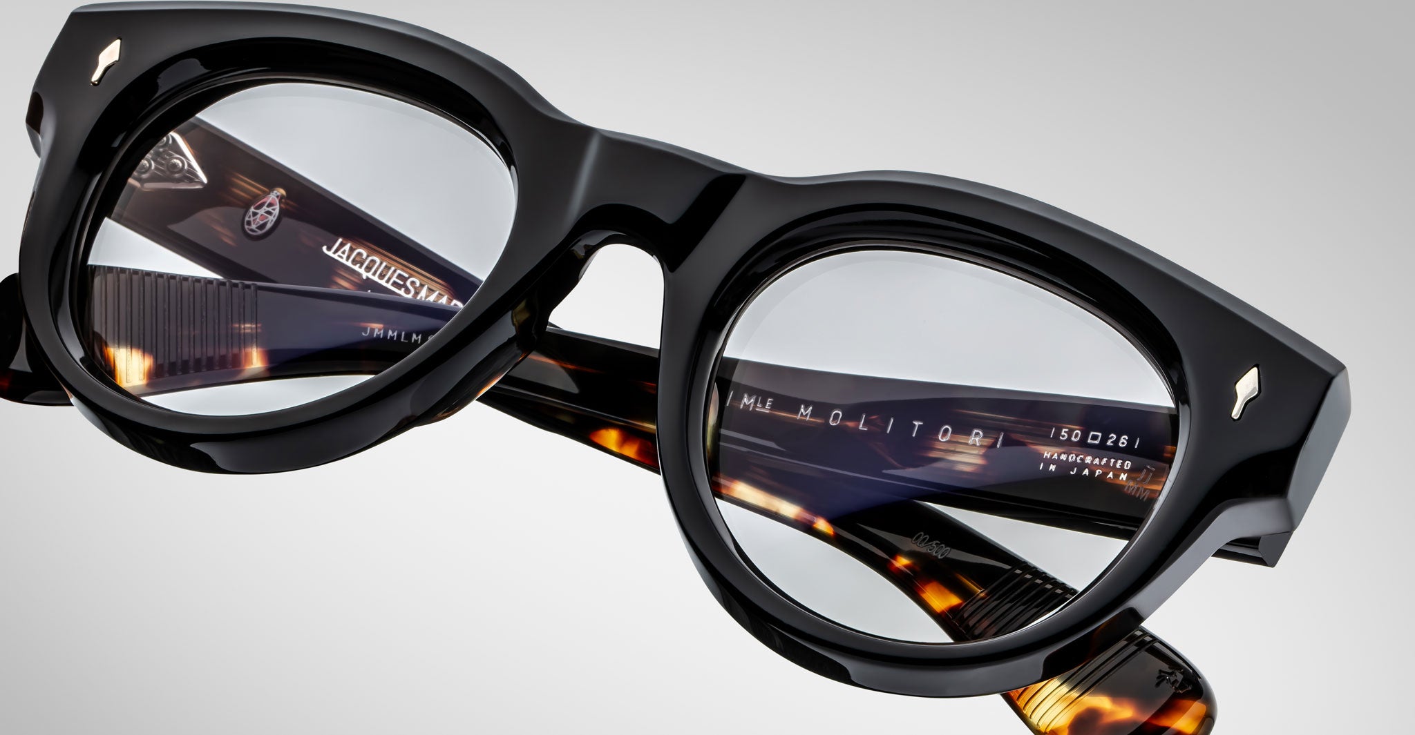Close-up of Jacques Marie Mage Molitor eyeglasses featuring round black and tortoiseshell frames. Brand markings and product details are visible on the inside of the temples. The frames rest on a white surface with a gradient background.