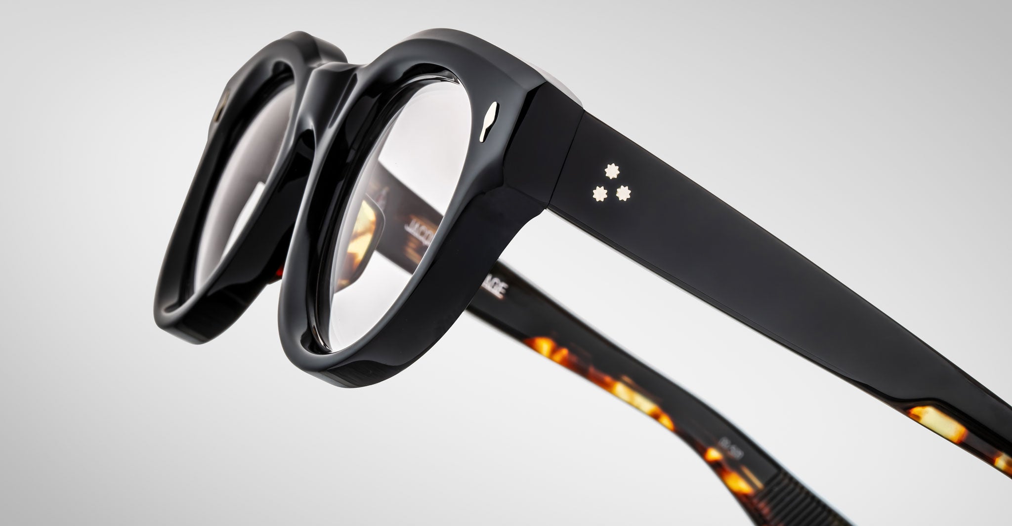 Close-up of Jacques Marie Mage Molitor eyeglasses in black with clear lenses, featuring two small star-shaped silver accents on the temples and patterned brown details on the inner arms.