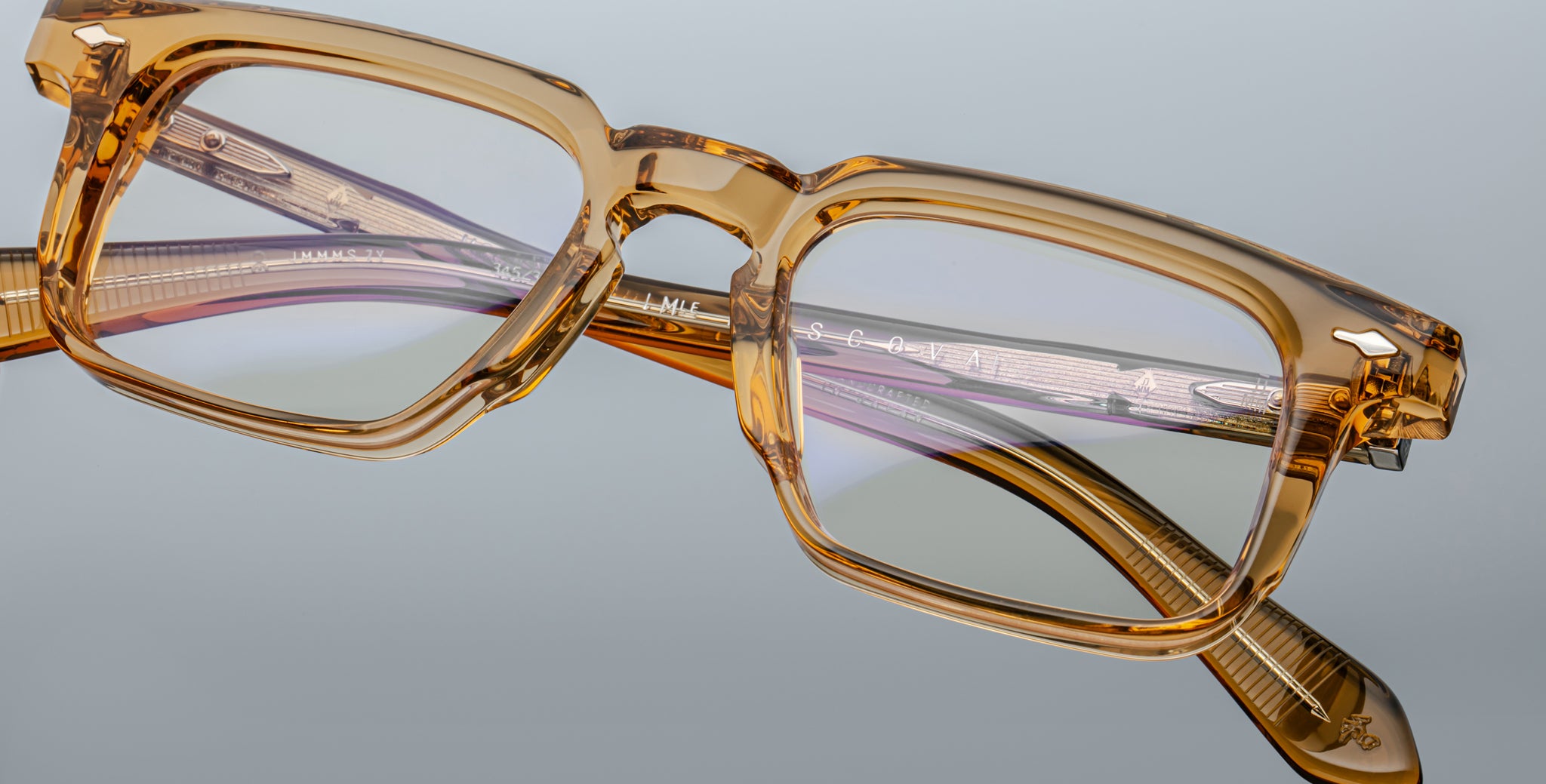 Close-up of Jacques Marie Mage Moscova eyeglasses, featuring thick rectangular translucent amber frames with visible hinges, engraved temples, and metal details, set against a plain gray background.