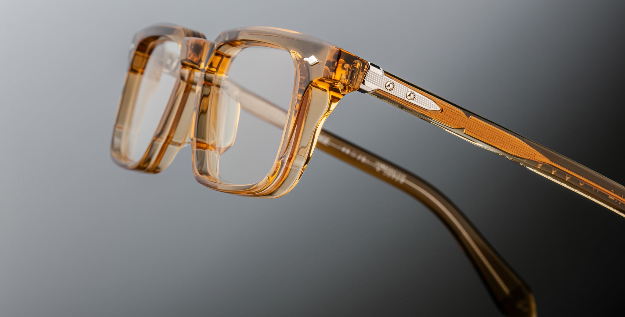 Close-up of Jacques Marie Mage Moscova eyeglasses featuring thick, transparent amber-colored frames and metal hinges, photographed against a gradient gray background.