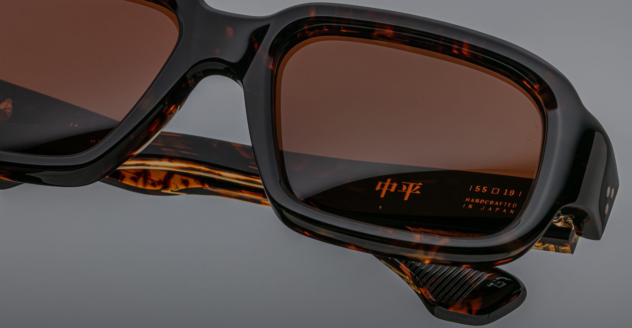 Close-up of Jacques Marie Mage Nakahira tortoiseshell sunglasses with brown lenses. The inner arm displays gold Japanese characters and HANDCRAFTED IN JAPAN on a plain gray background.