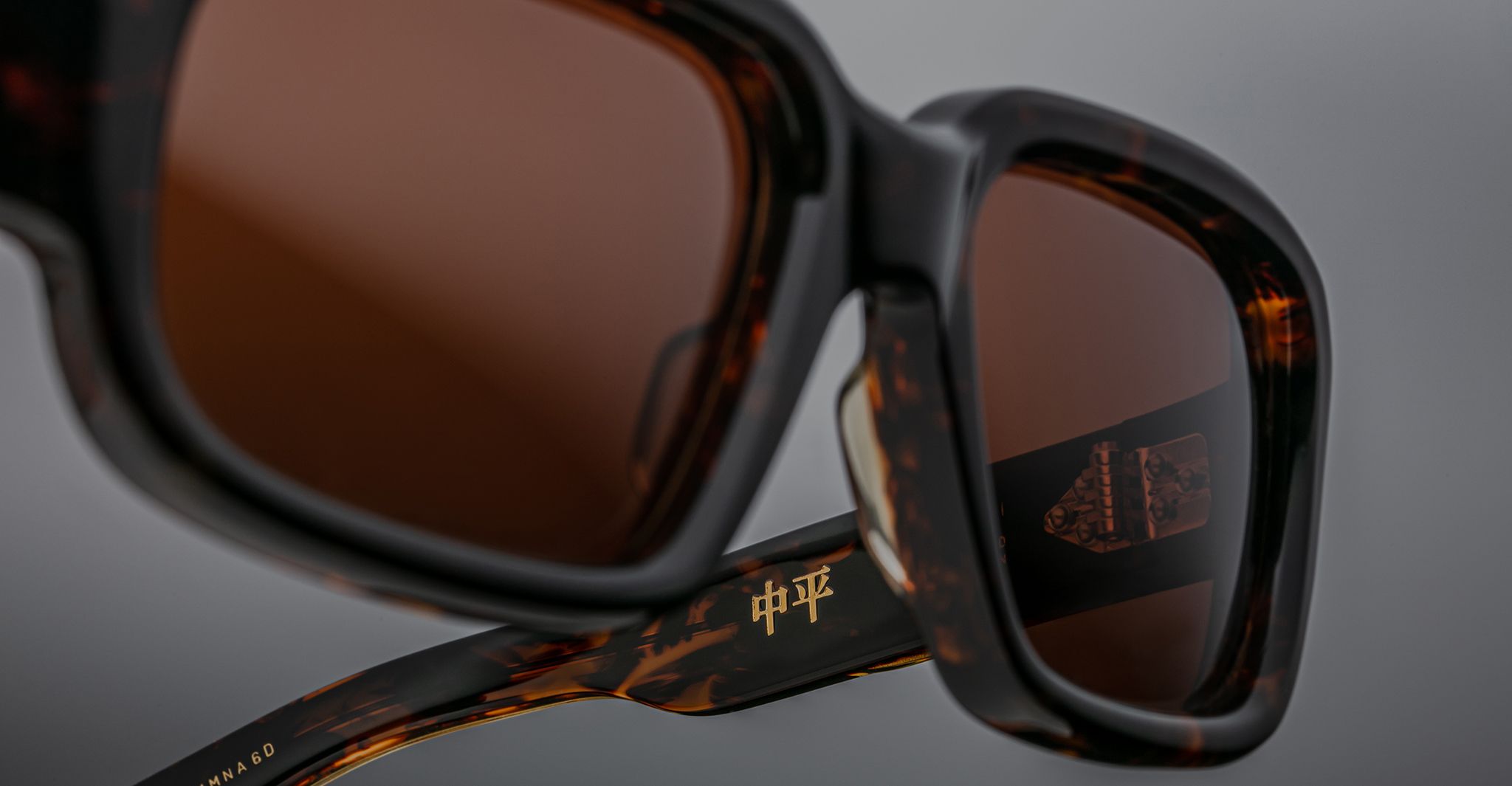 Close-up of Jacques Marie Mage Nakahira tortoiseshell sunglasses with brown lenses, featuring gold Chinese characters on the inner arm, set against a neutral gray background.