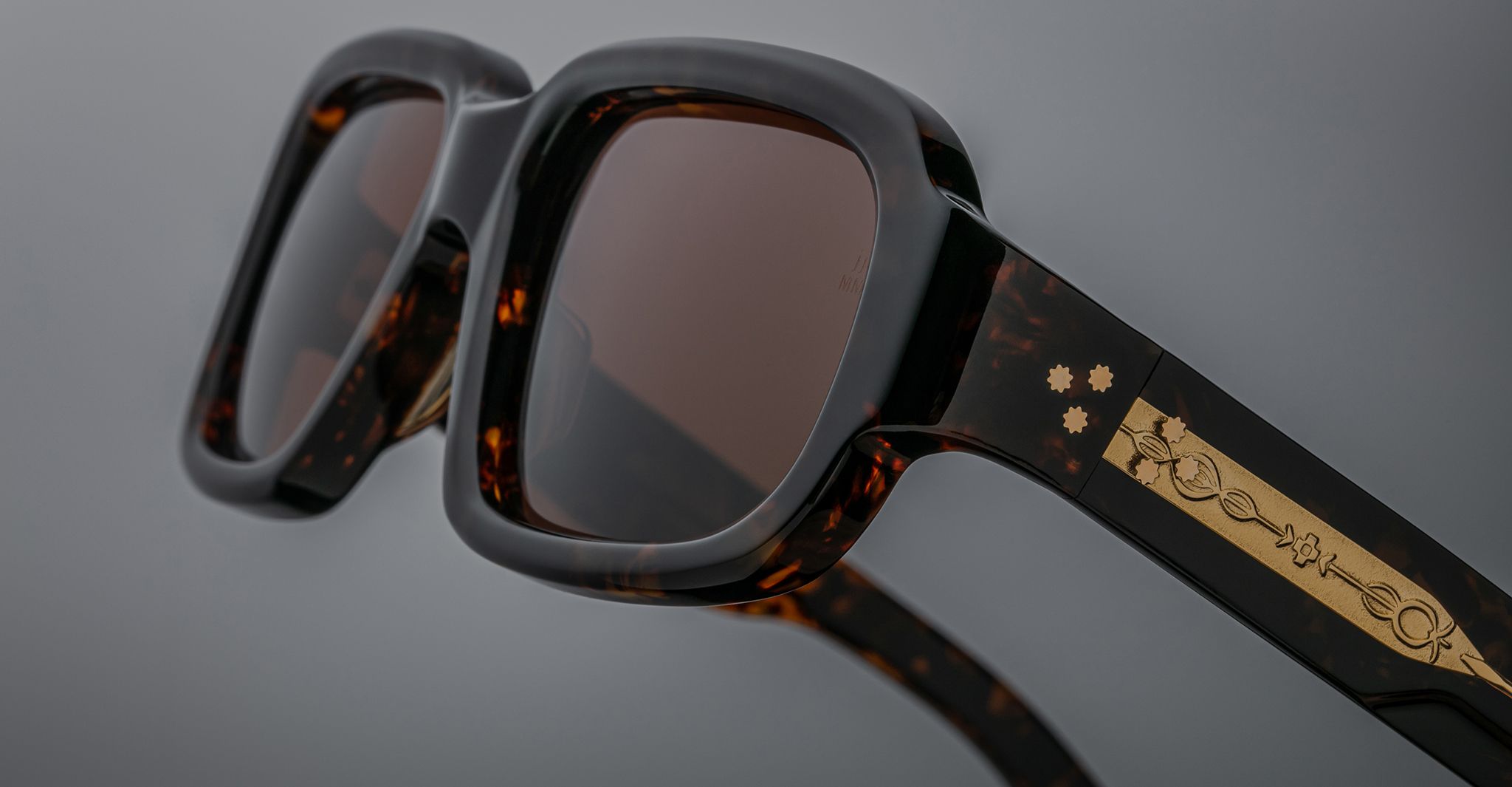 Close-up of Jacques Marie Mage Nakahira sunglasses—stylish, square tortoiseshell frames with dark lenses and a gold decorative pattern on the temple—set against a neutral gray background.