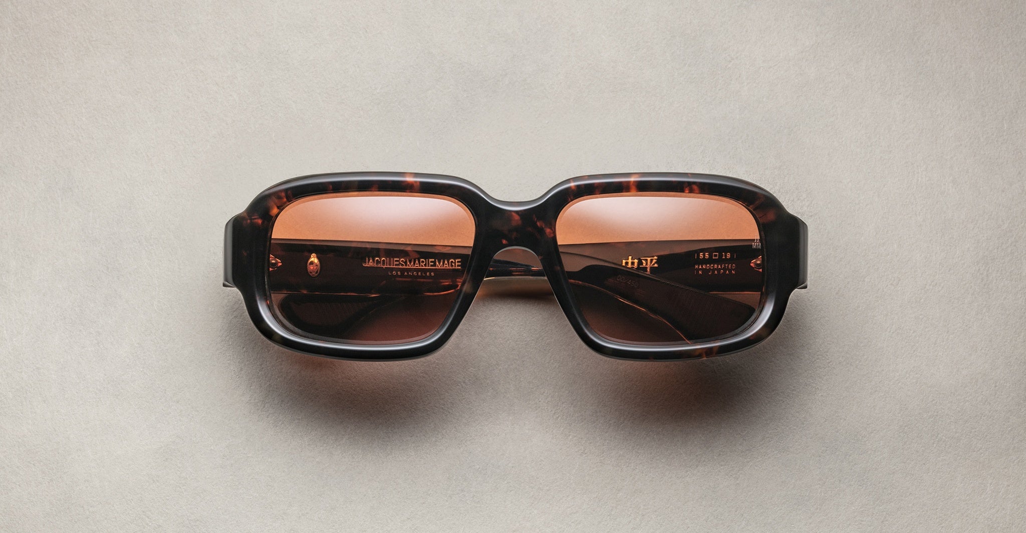 Jacques Marie Mage Nakahira rectangular tortoiseshell sunglasses with thick frames and brown tinted lenses, shown on a light beige surface.
