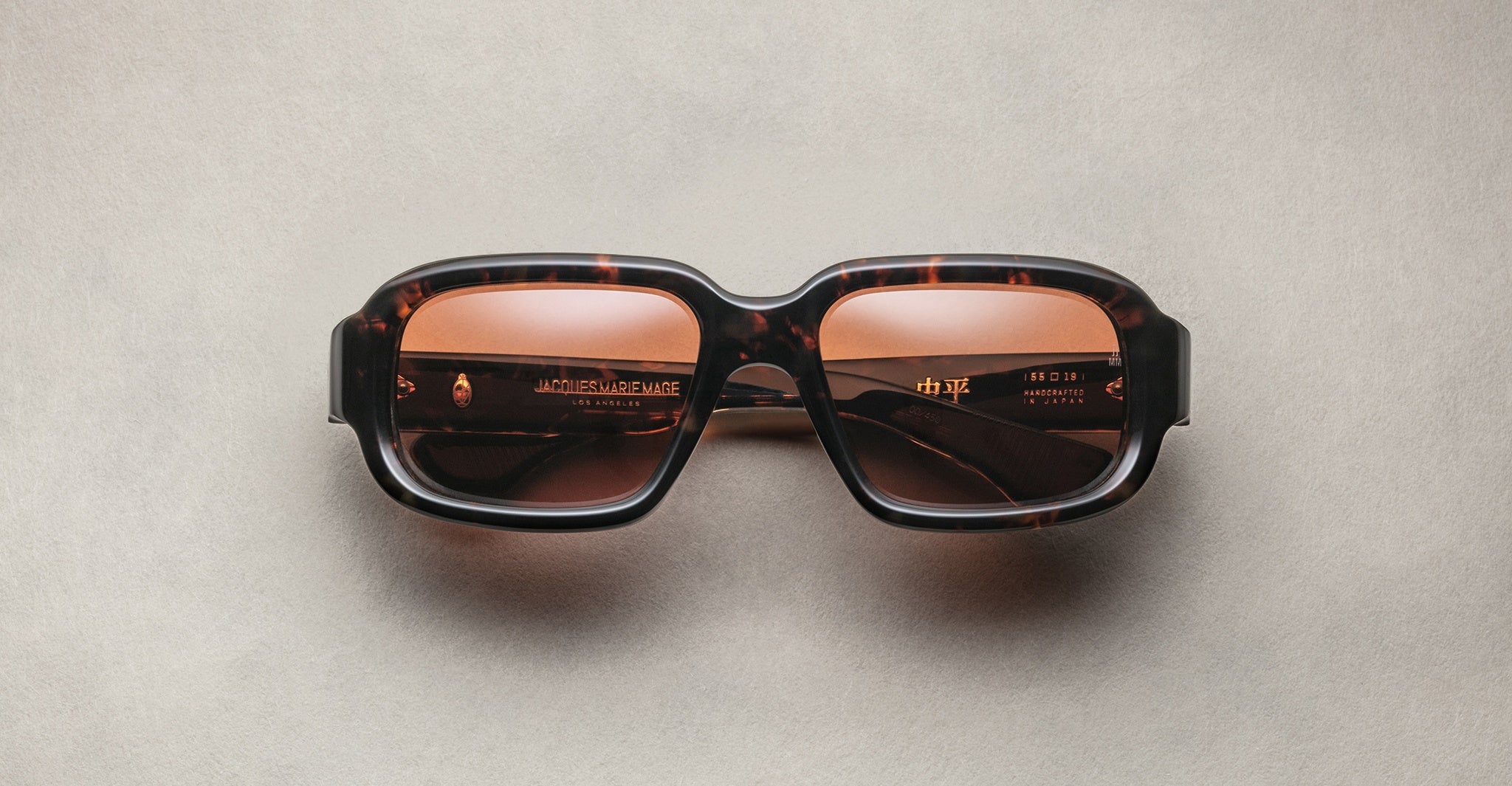 Jacques Marie Mage Nakahira rectangular tortoiseshell sunglasses with thick frames and brown tinted lenses, shown on a light beige surface.
