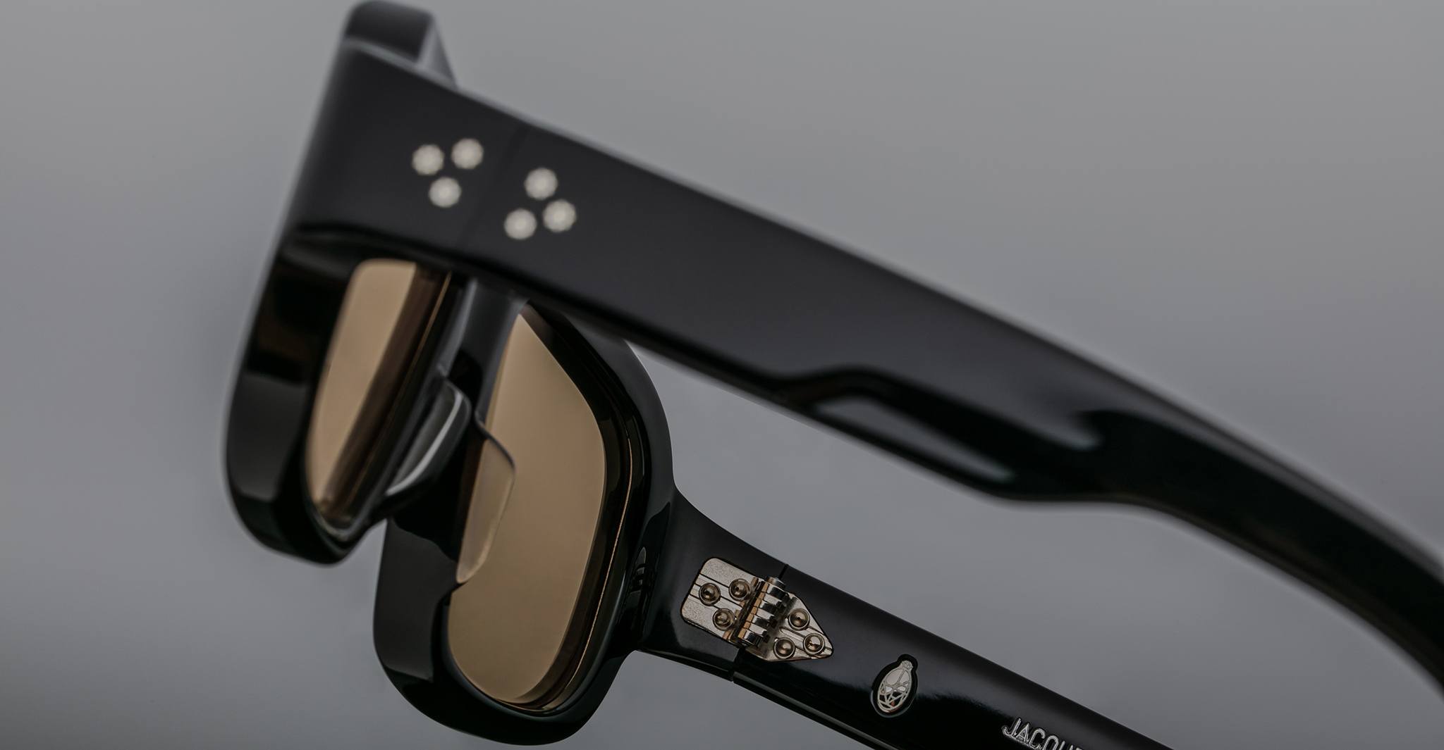 A close-up of the temple arms of Jacques Marie Mage Nakahira black sunglasses, featuring metal hinge detailing and three small circular dots on the side, set against a plain grey background.