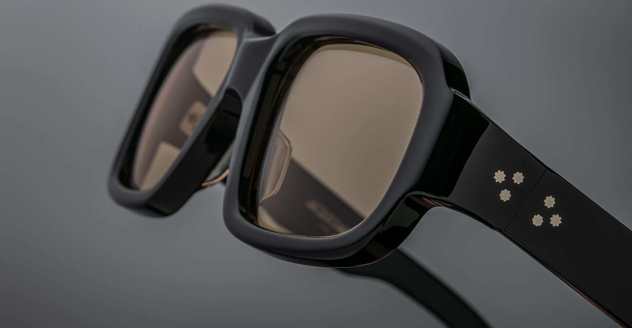 Close-up of Jacques Marie Mage Nakahira black rectangular sunglasses with gold star-shaped accents on the arms, set against a neutral gray background.