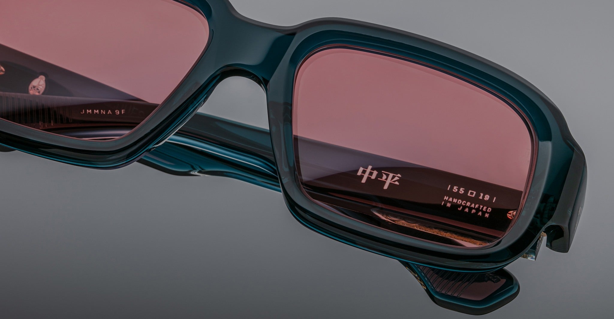 Close-up of Jacques Marie Mage Nakahira sunglasses with thick, dark green frames and rose-tinted lenses; “Handcrafted in Japan” and Japanese characters are visible on the inner arm.