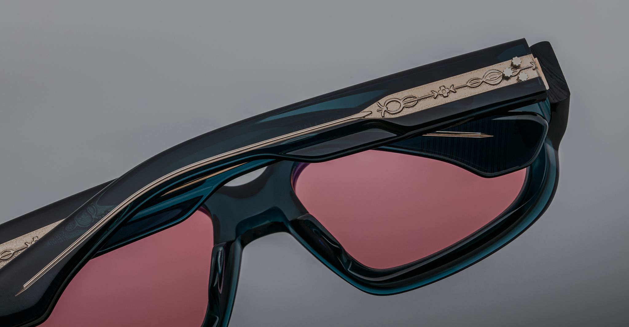Close-up of Jacques Marie Mage Nakahira sunglasses featuring dark blue-green frames, pink-tinted lenses, and an ornate metallic temple design, set against a neutral gray background.