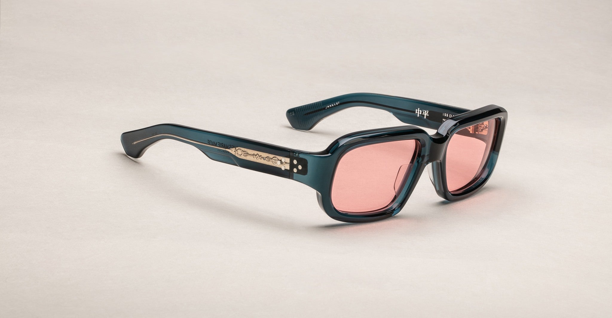 The Jacques Marie Mage Nakahira sunglasses feature square-shaped, thick dark teal frames with pink-tinted lenses and gold temple details, displayed on an off-white background.