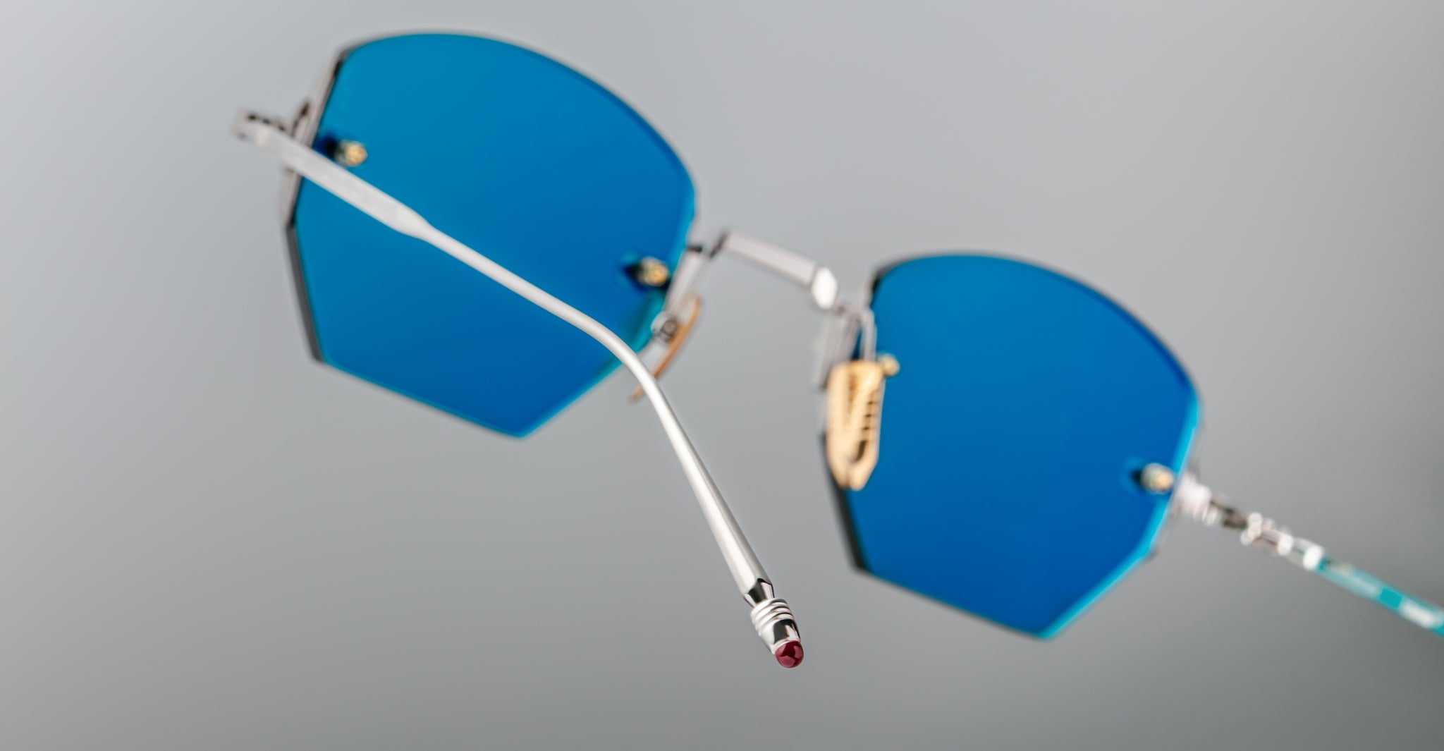 Close-up of Jacques Marie Mage Oatman hexagonal rimless sunglasses featuring geometric blue-tinted lenses and thin silver metal frames, highlighting exquisite handmade Japanese craftsmanship against a plain gray background.