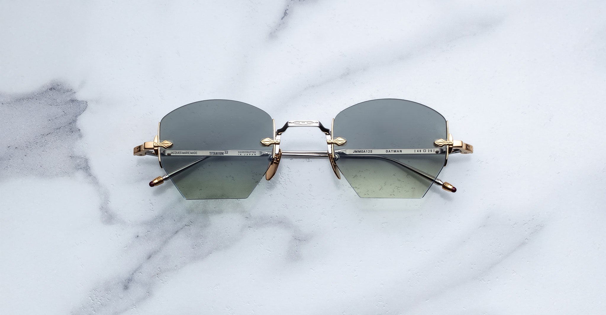 A pair of Jacques Marie Mage Oatman hexagonal rimless sunglasses with geometric gray lenses and gold metal temples rests on a white marble surface.