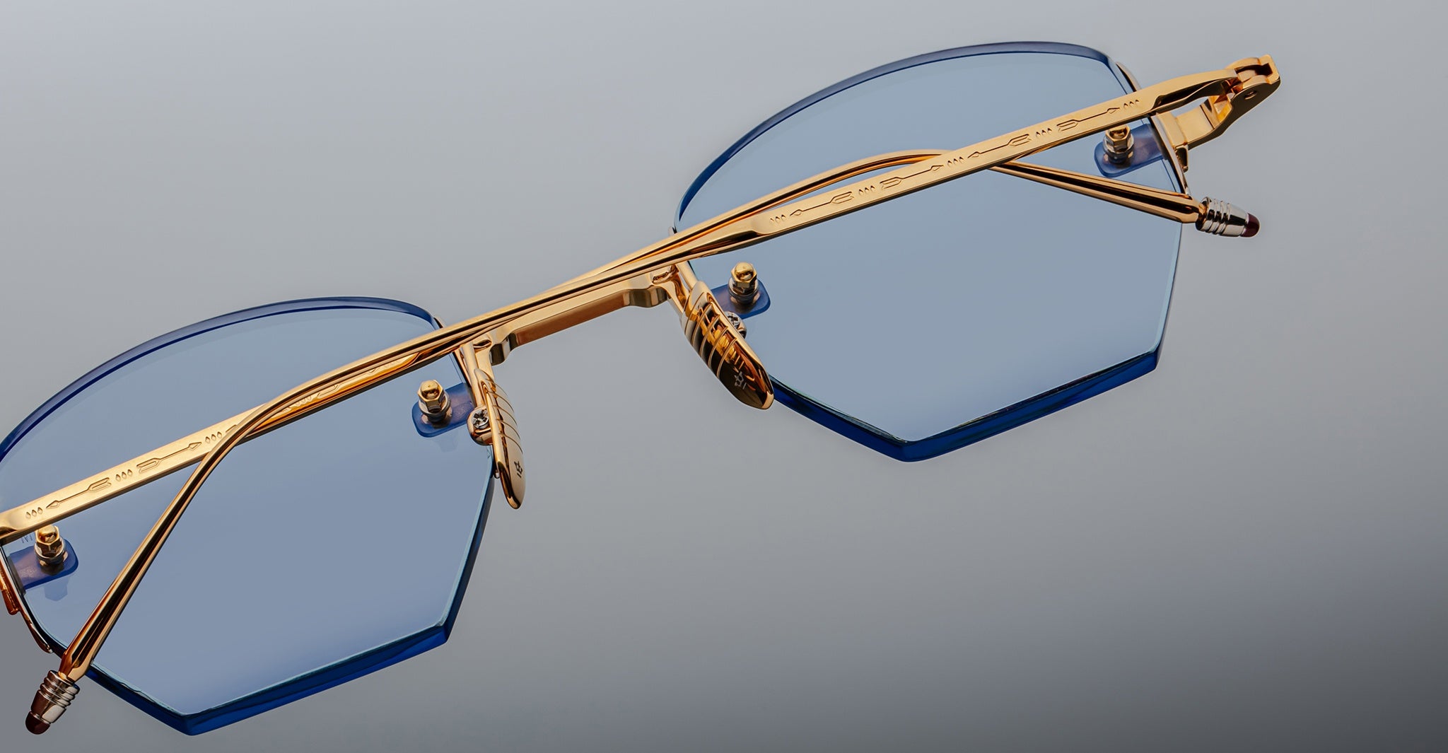 Close-up of Jacques Marie Mage Oatman hexagonal eyeglasses with blue-tinted lenses and a gold metal frame, shown against a neutral background—a striking example of handmade Japanese eyewear from Jacques Marie Mage.