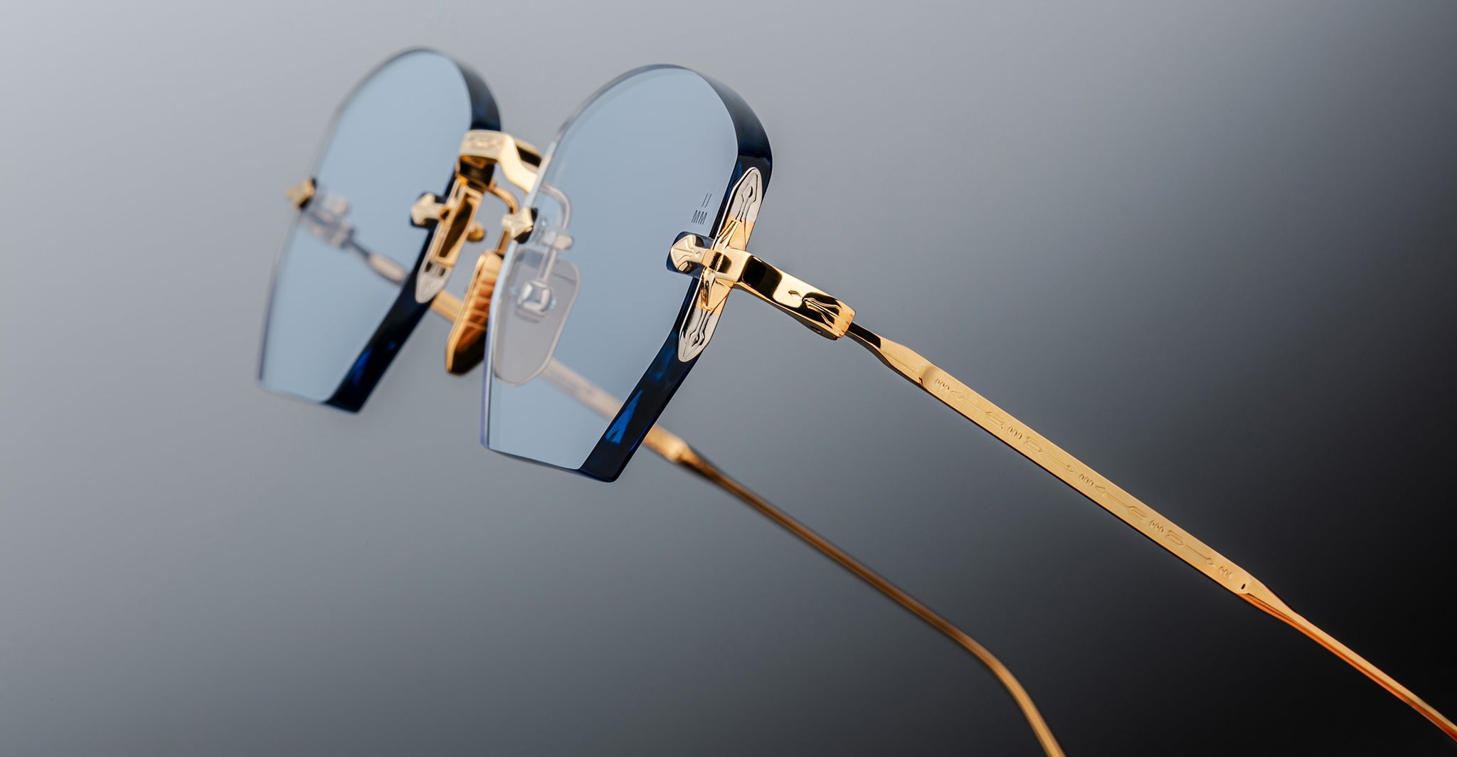 A close-up of Jacques Marie Mage Oatman hexagonal rimless sunglasses, featuring blue-tinted lenses and gold metal temples by Jacques Marie Mage, set against a gradient gray background.
