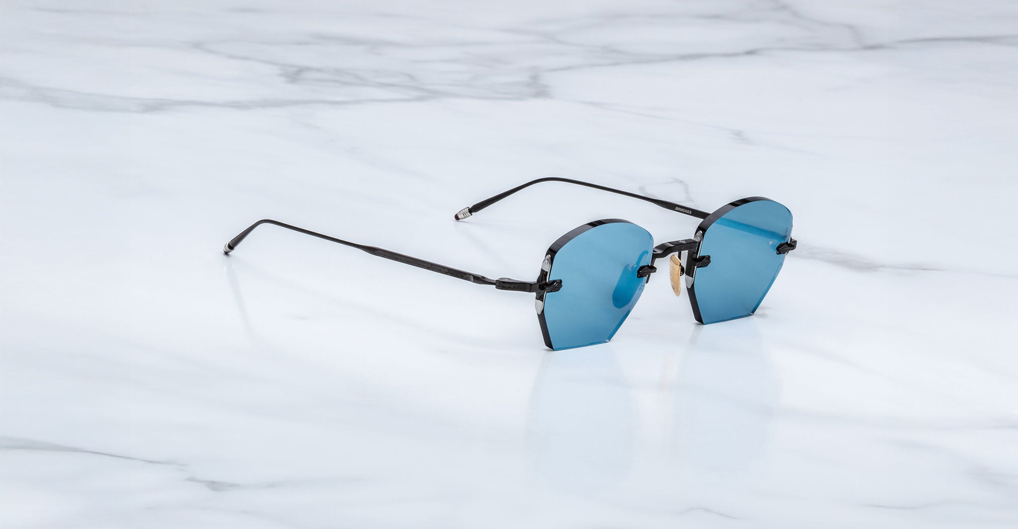 The Jacques Marie Mage Oatman, handmade Japanese hexagonal rimless sunglasses with blue-tinted lenses and thin black arms, is displayed on a white marble surface.