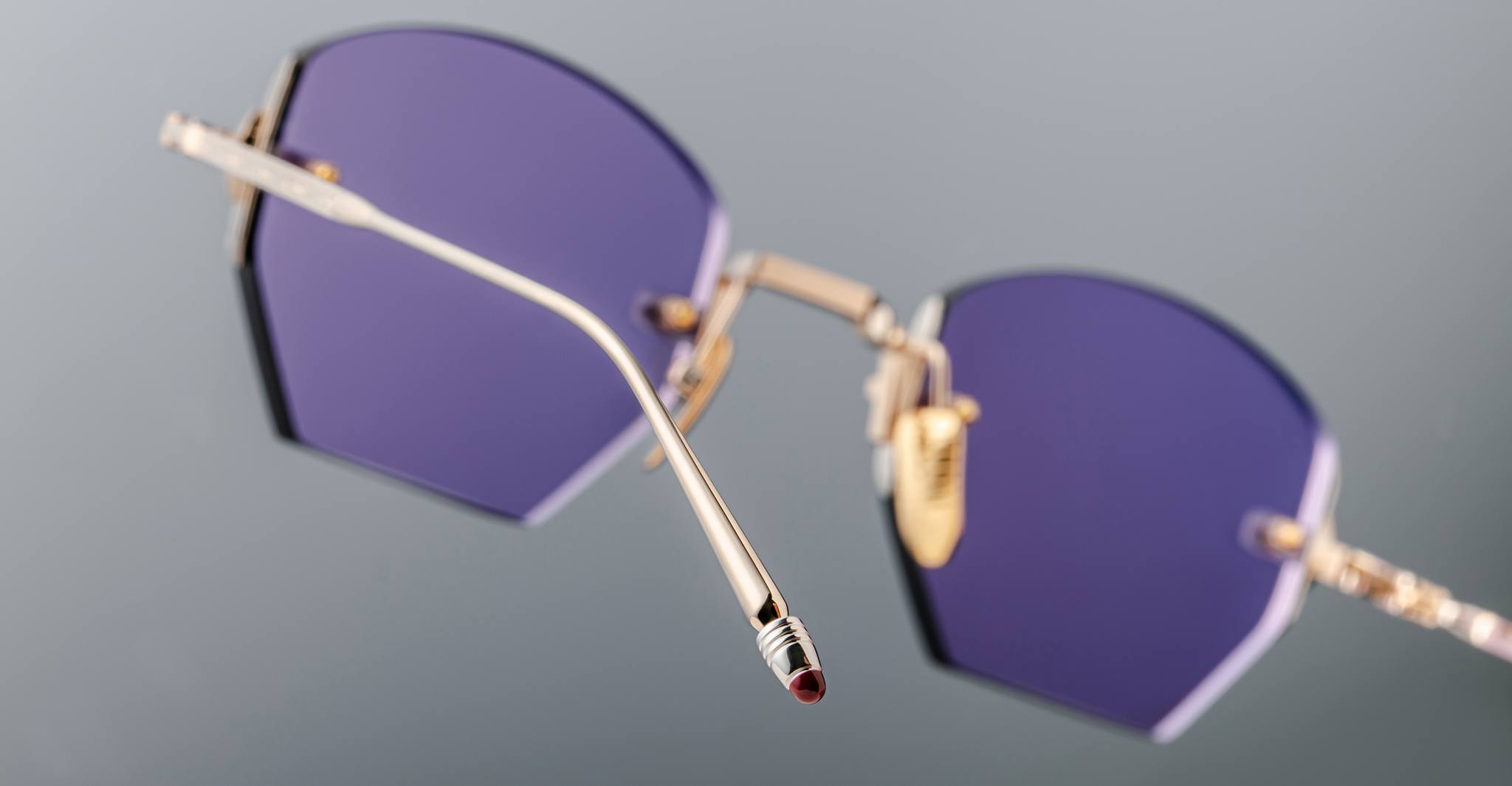 Close-up of Jacques Marie Mage Oatman: handmade Japanese gold-framed, hexagonal rimless sunglasses featuring geometric purple-tinted lenses by Jacques Marie Mage, set against a plain grey background.