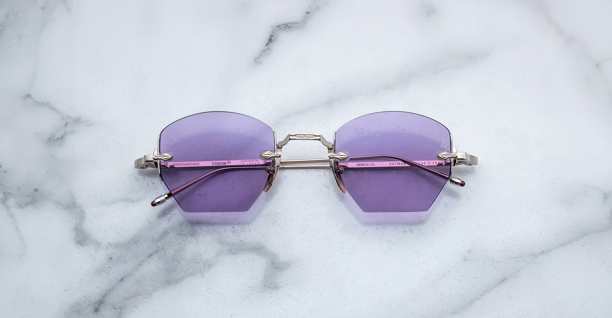 Jacques Marie Mage Oatman sunglasses by Jacques Marie Mage feature hexagonal rimless frames, purple lenses, and silver arms inspired by Japanese craftsmanship, shown elegantly on a white marble surface.