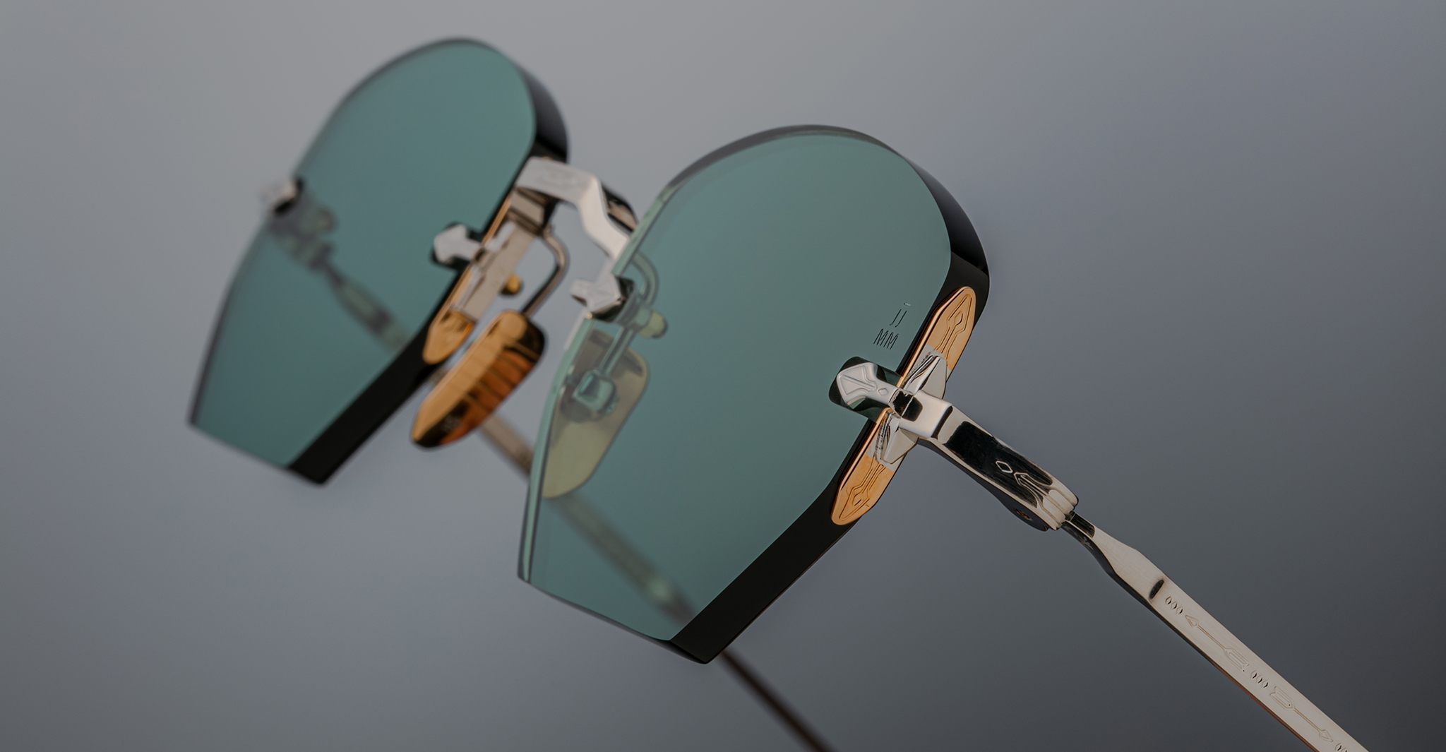 Close-up of Jacques Marie Mage Oatman eyewear: handmade hexagonal rimless sunglasses with green lenses, silver metal frames, and wooden temples by Jacques Marie Mage, displayed against a neutral background.