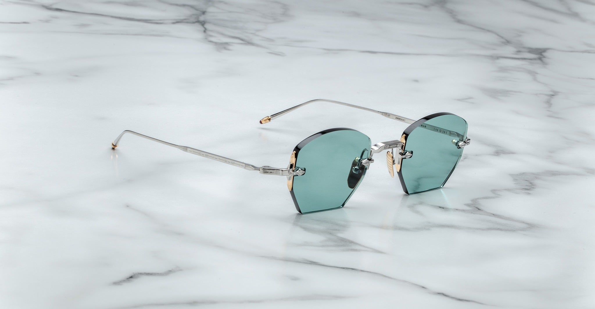 A pair of Jacques Marie Mage Oatman round rimless sunglasses with blue-green tinted lenses and thin metal arms rests on a white marble surface.