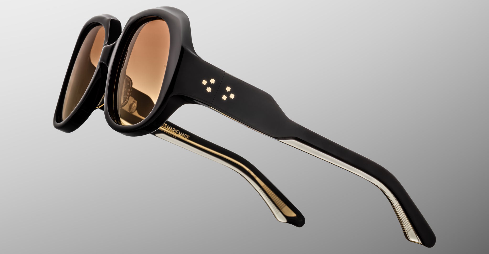 The Jacques Marie Mage Ocean Child sunglasses feature thick black frames, brown-tinted lenses, and four small star-shaped metallic accents on the temples, displayed against a plain gray backdrop.