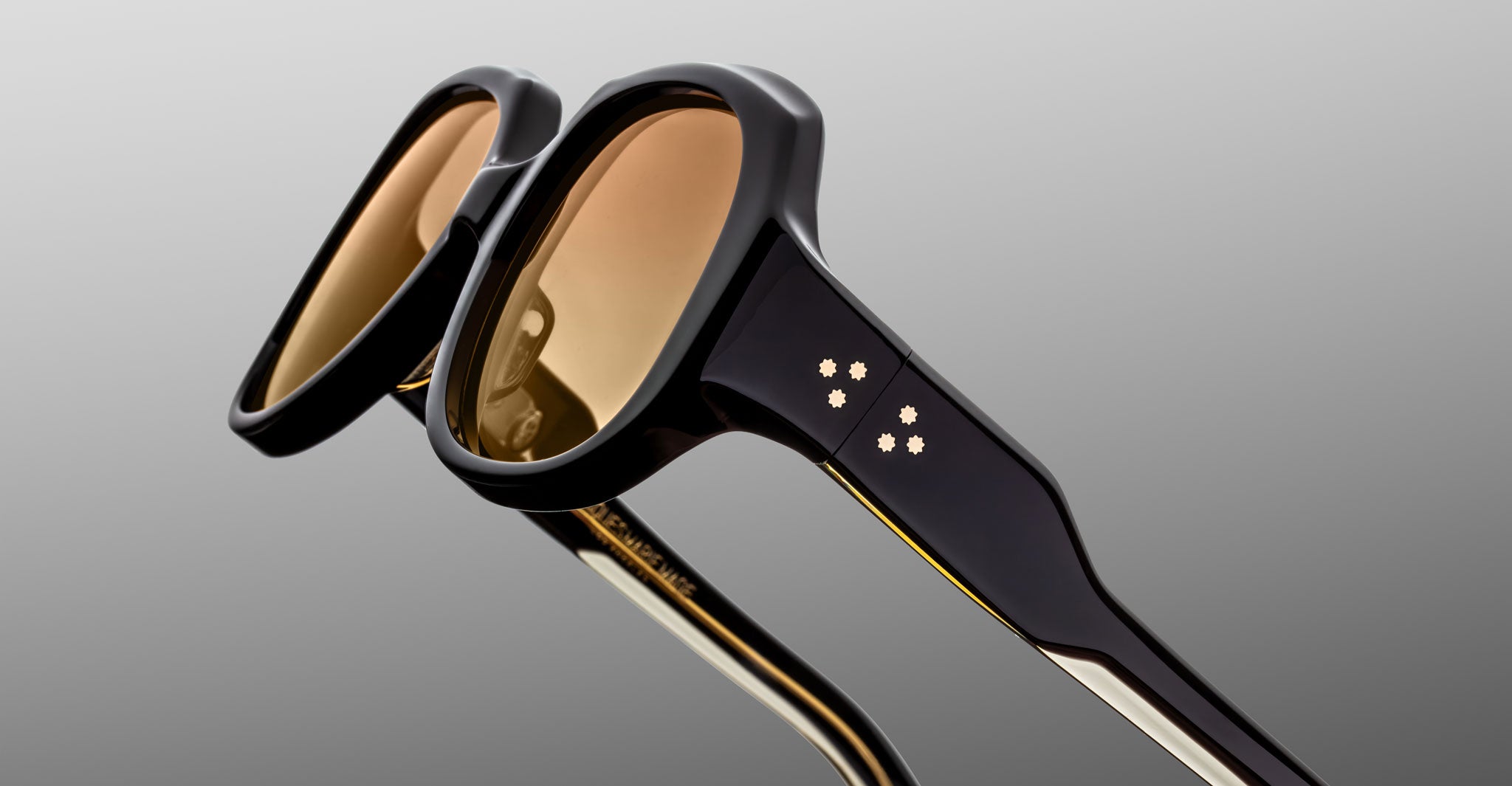 Close-up of Jacques Marie Mage Ocean Child sunglasses with thick black frames, brown-tinted lenses, and gold star-shaped accents on the temples, set against a gradient gray background.