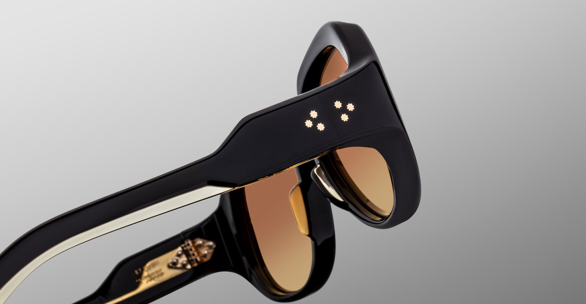 Close-up of Jacques Marie Mage Ocean Child sunglasses in black with brown lenses, showcasing four small star-shaped accents on the temple against a light gray background.