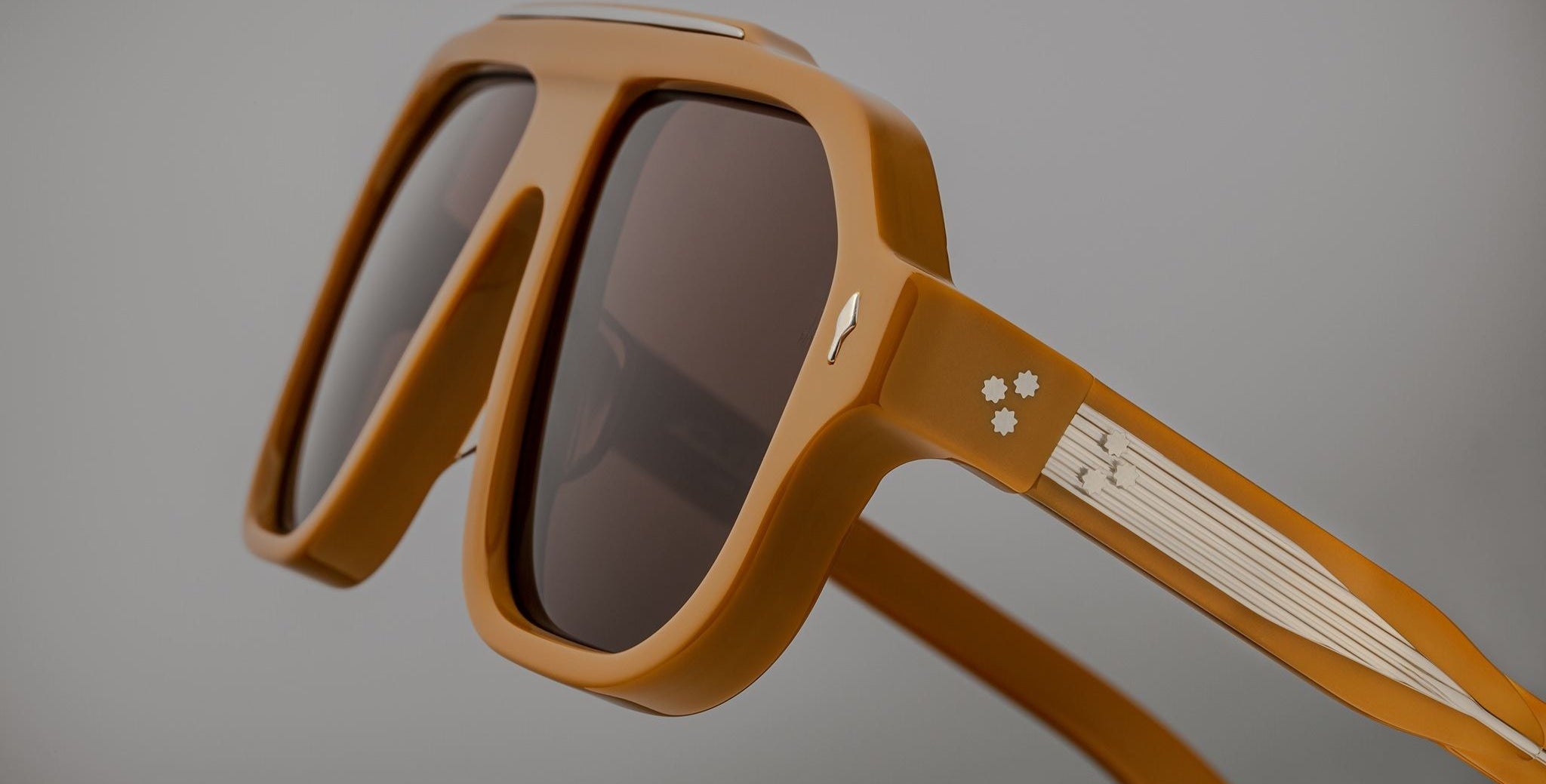 Close-up of Jacques Marie Mage Octavian oversized square sunglasses with a thick matte brown frame and dark lenses. The temples have a white accent with 3 small star rivets near the hinge, set against a plain gray background.