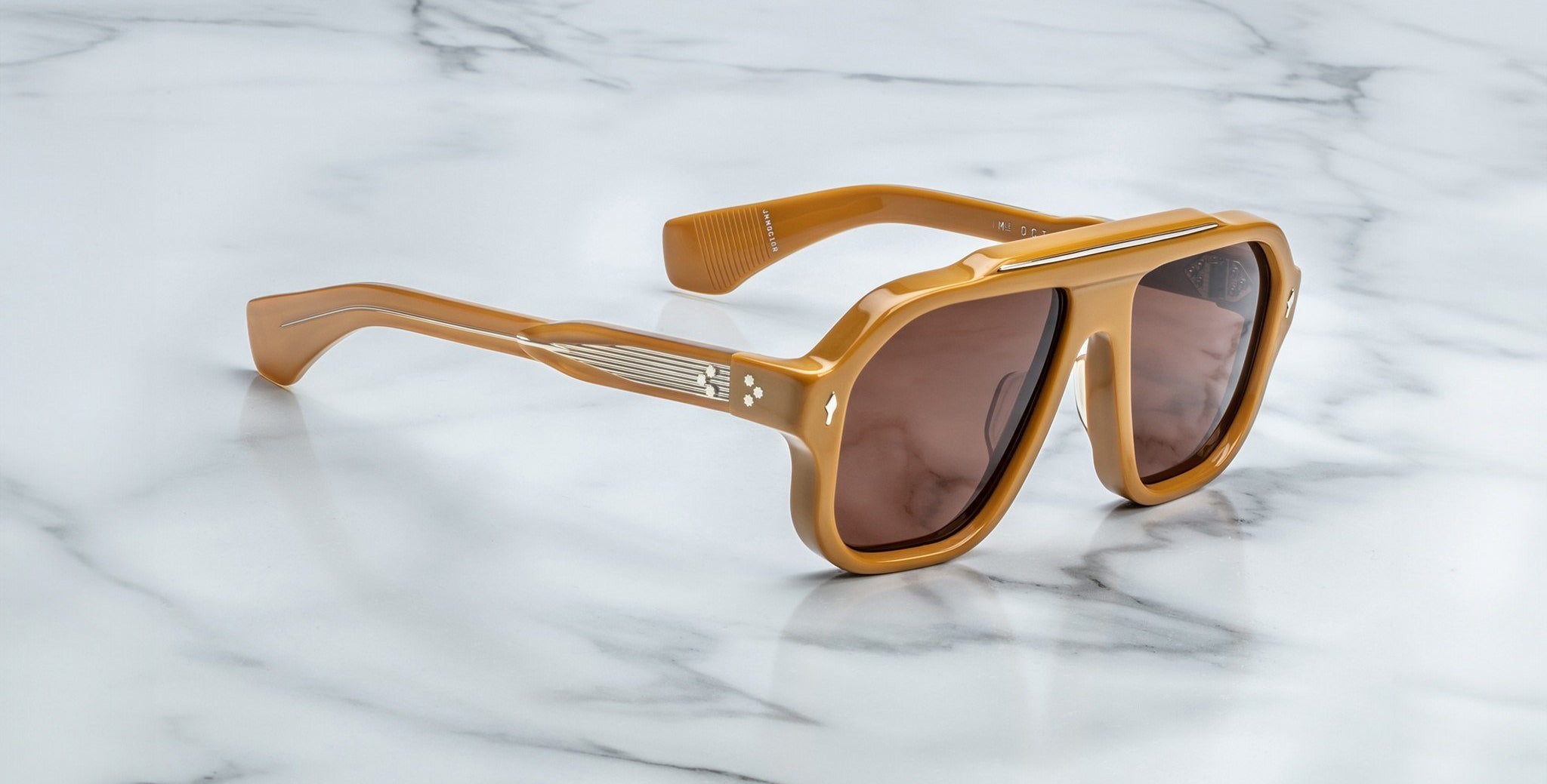 The Jacques Marie Mage Octavian sunglasses, featuring oversized brown frames with tinted lenses, thick angular design, and metallic accents on the arms, rest on a polished white marble surface.