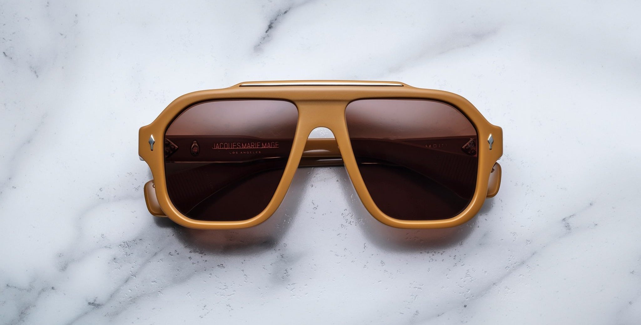 The Jacques Marie Mage Octavian sunglasses, featuring brown frames and dark lenses, rest face up on a white marble surface.