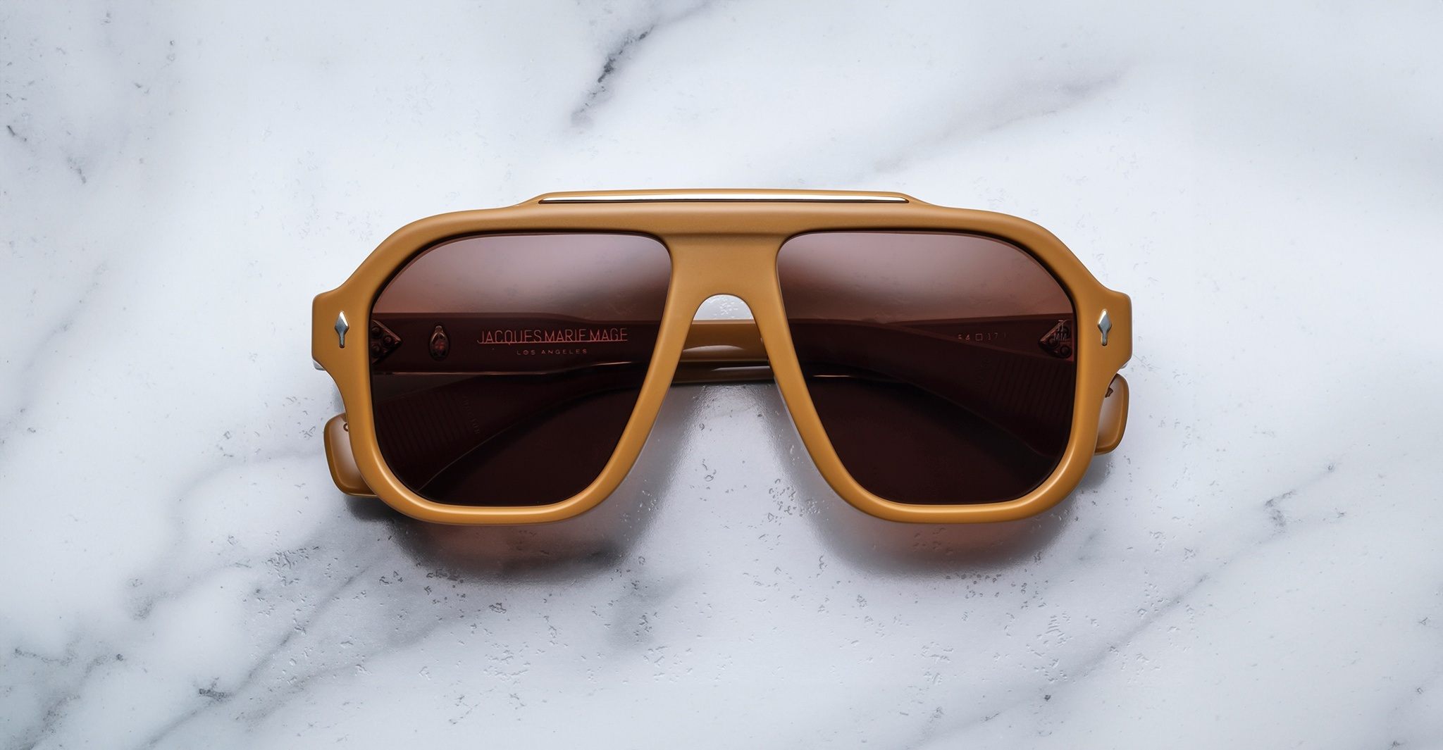 The Jacques Marie Mage Octavian sunglasses, featuring brown frames and dark lenses, rest face up on a white marble surface.